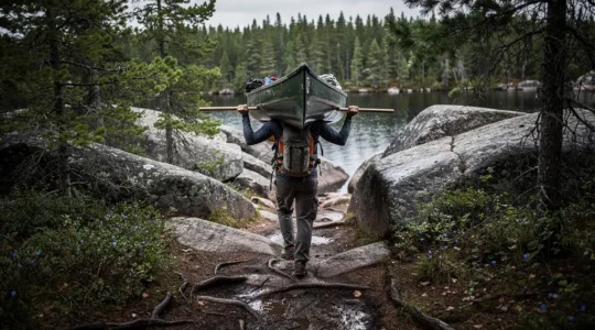 A first-time canoe tripper carries a loaded canoe on a portage yoke along a rocky Canadian Shield trail between two lakes.